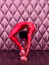 Wearing the Showgirl Gymnastic Costume—a custom-made, red sequin bodysuit—a performer executes an extreme backbend atop wooden blocks in front of a quilted wall.