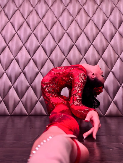 A dancer in a custom-made red sheer Showgirl Gymnastic Costume, a sequin bodysuit for women or men, bends backward on a wooden floor against a quilted wall.