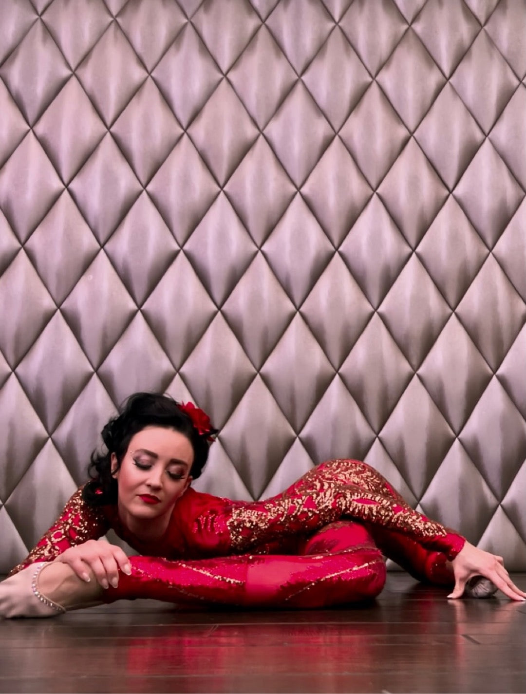 A woman wearing the custom-made, red Showgirl Gymnastic Sequin Bodysuit performs a stunning contortion pose on a wooden floor with a patterned wall.