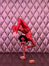 A performer, dressed in a custom Showgirl Gymnastic Costume— a red sequin bodysuit made to order— balances on one hand atop a wooden block, set against a quilted backdrop.