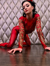 A woman wears the Showgirl Gymnastic Costume—a custom-made, red sequin bodysuit—posing on the floor on her hands and knees and gazing at the camera, with a quilted wall as a backdrop. Her shimmering jumpsuit catches the light, highlighting its tailored design for dance competitions or special occasions.