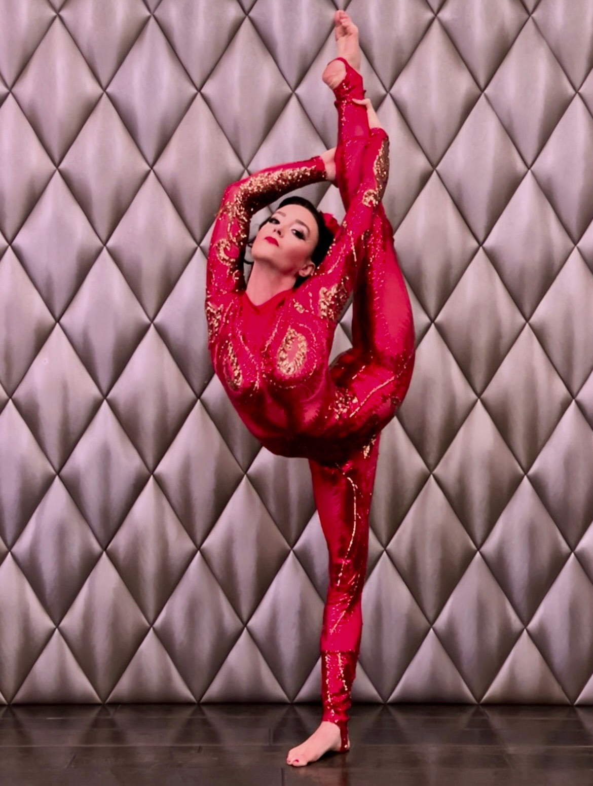 Wearing a custom-made Showgirl Gymnastic Costume—a red sheer sequin bodysuit designed for women or men—a woman performs a standing split against a padded wall background.