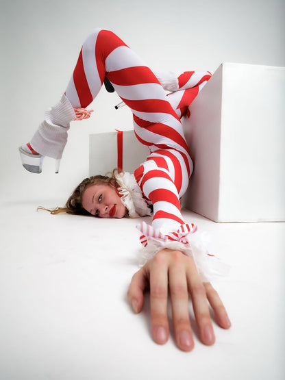 A woman in the Candy Girl Red and White Stripes Catsuit with full sleeves and a turtleneck poses contorted on the floor, reaching toward the camera in her striking red and white bodysuit.
