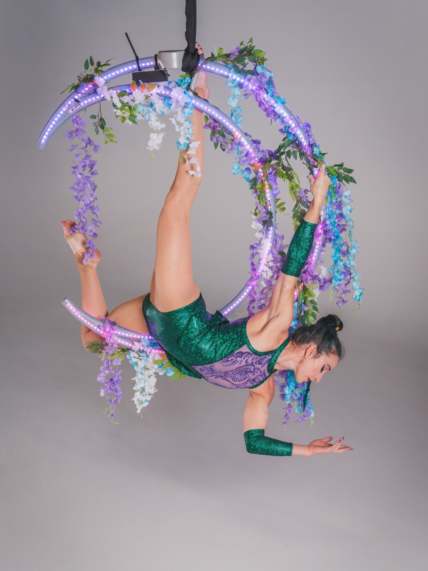 An aerial artist poses gracefully on a floral-lit hoop, wearing the Shattered Emerald Skater Leotard with halter neck and sheer sequin details.
