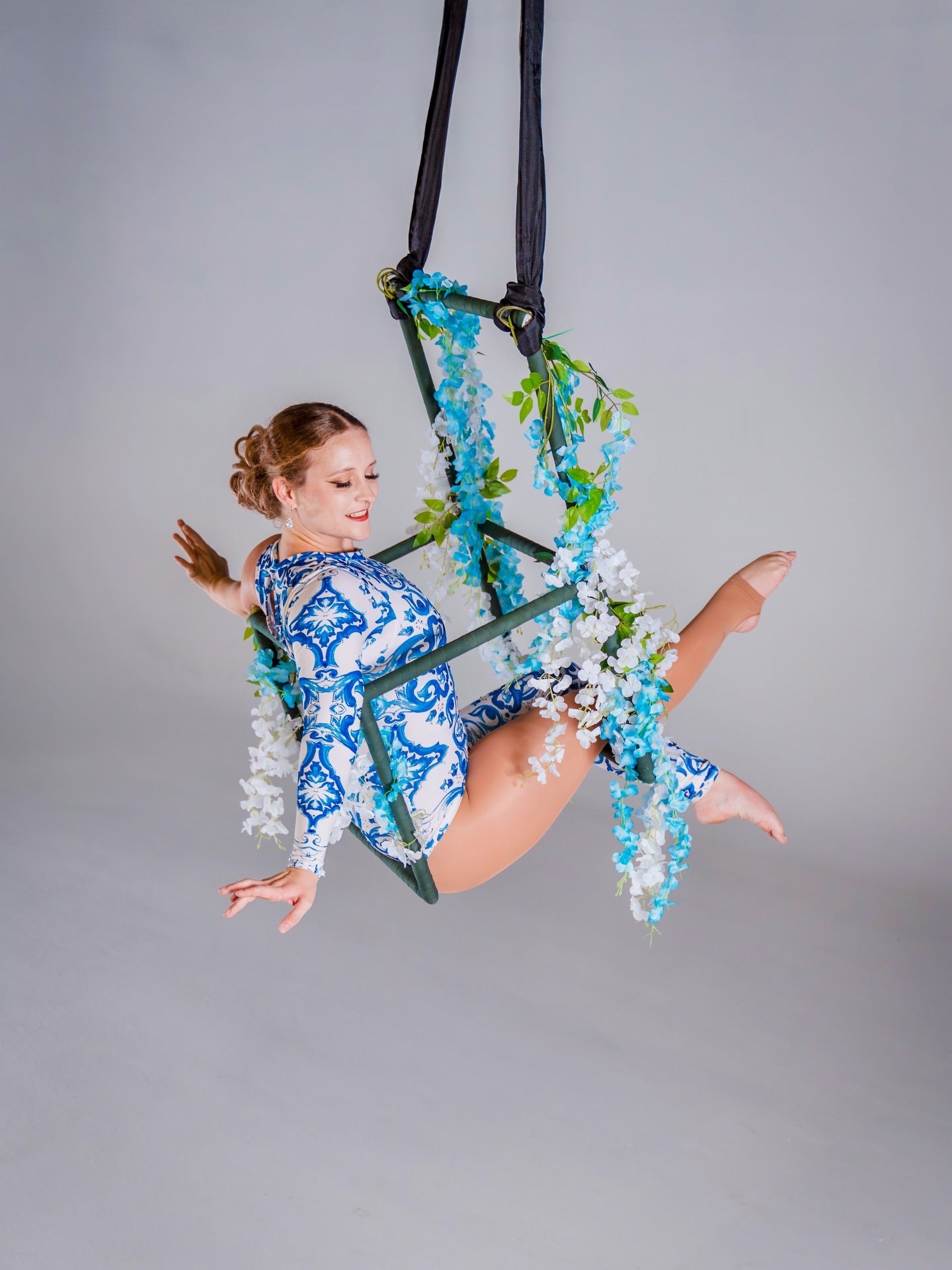 Woman wears an Asymmetrical Catsuit for Performers—blue &amp; white porcelain print, one-sleeve, one-leg stretch spandex bodysuit with baroque scroll pattern—posing on a floral hoop against a plain backdrop.