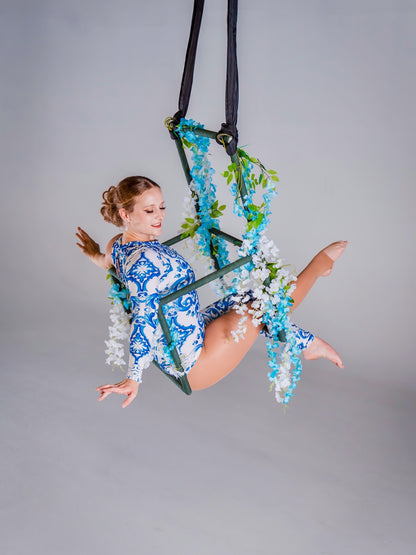 Woman wears an Asymmetrical Catsuit for Performers—blue &amp; white porcelain print, one-sleeve, one-leg stretch spandex bodysuit with baroque scroll pattern—posing on a floral hoop against a plain backdrop.
