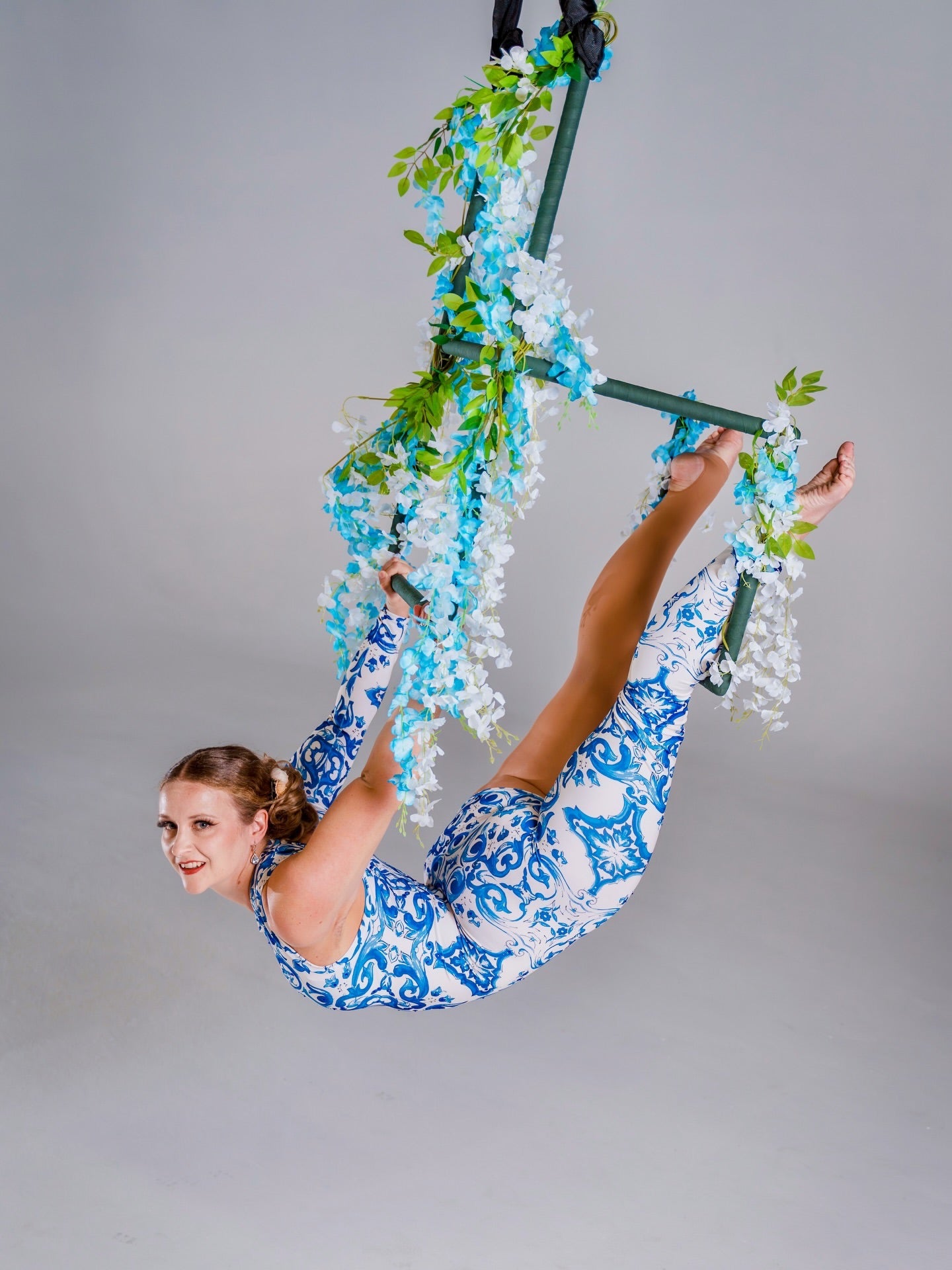 A woman wears the Asymmetrical Catsuit for Performers—a blue &amp; white porcelain print, one-sleeve, one-leg spandex jumpsuit with a baroque scroll pattern—while performing on a floral aerial hoop against a plain background.