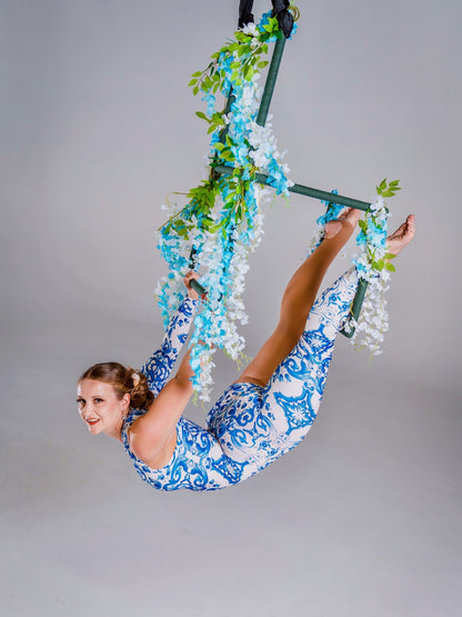 A woman wears the Asymmetrical Catsuit for Performers—a blue &amp; white porcelain print, one-sleeve, one-leg spandex jumpsuit with a baroque scroll pattern—while performing on a floral aerial hoop against a plain background.