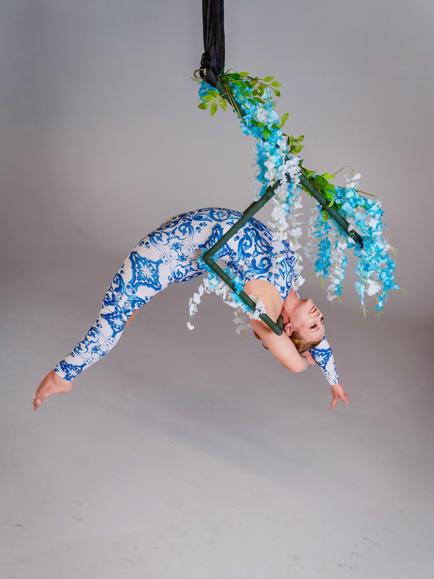 Aerial performer in an asymmetrical catsuit featuring a blue &amp; white porcelain baroque scroll print hangs from a hoop decorated with blue and white flowers. This stretch spandex unitard is perfect for dance and events.