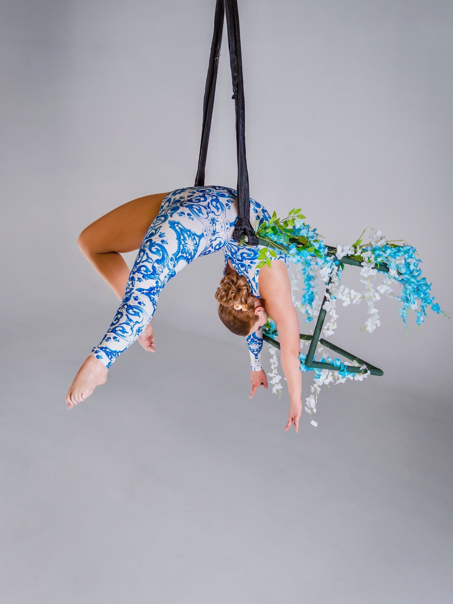 An aerial artist in a blue &amp; white porcelain print asymmetrical catsuit performs suspended upside down on a floral-decorated hoop. The stretch spandex bodysuit features one sleeve, one leg, and a baroque scroll pattern—perfect for dance &amp; events.