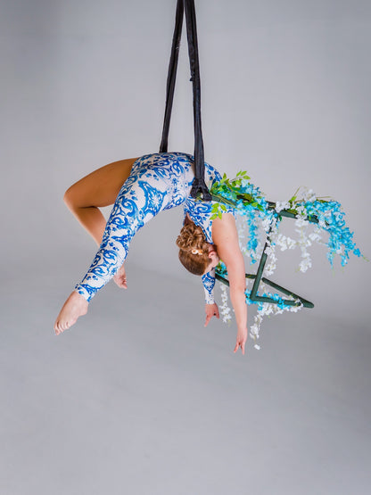 An aerial artist in a blue &amp; white porcelain print asymmetrical catsuit performs suspended upside down on a floral-decorated hoop. The stretch spandex bodysuit features one sleeve, one leg, and a baroque scroll pattern—perfect for dance &amp; events.