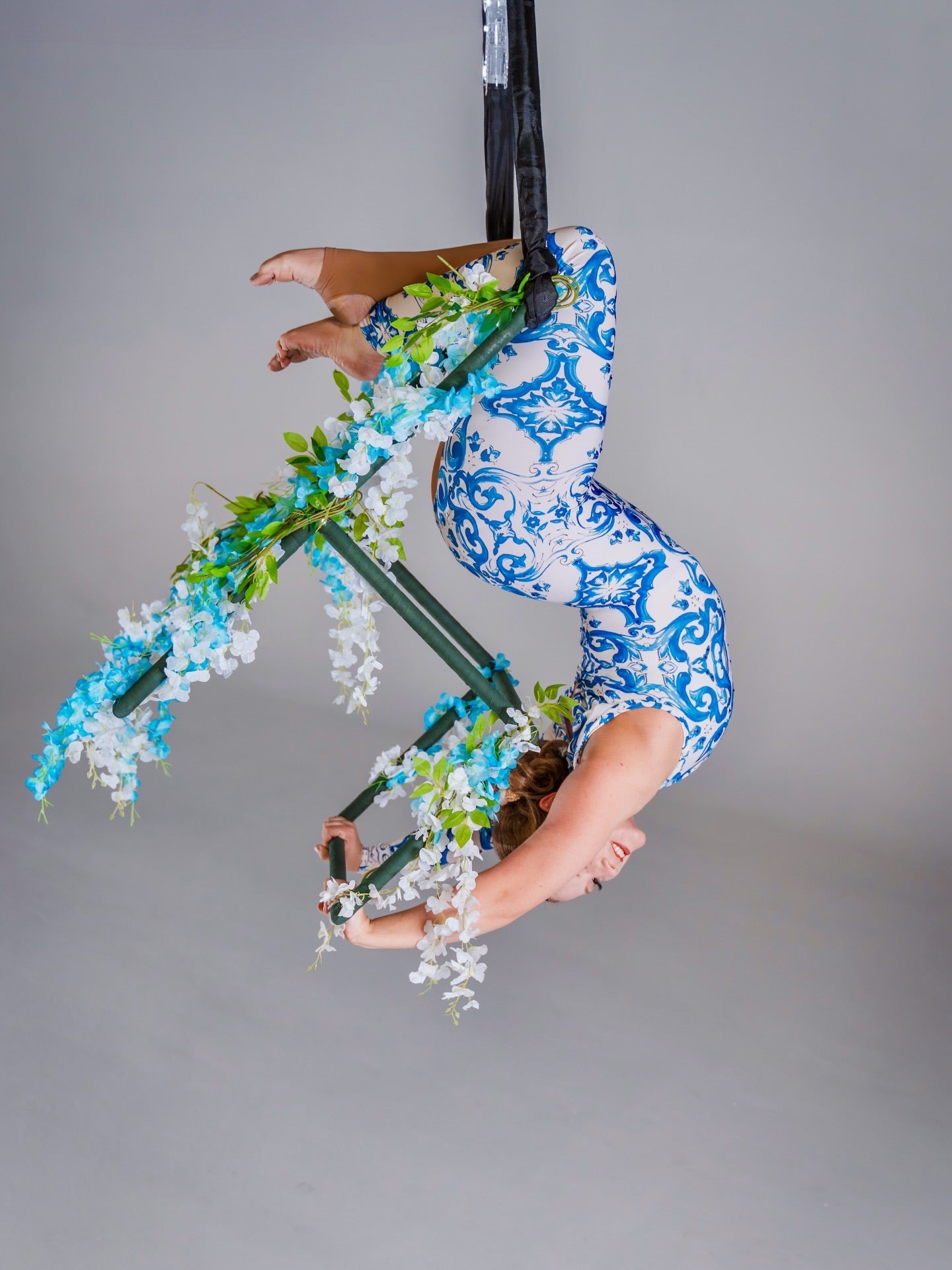 A woman in an Asymmetrical Catsuit for Performers, featuring a blue &amp; white porcelain print and baroque scroll pattern, performs aerial acrobatics on a hoop decorated with flowers.