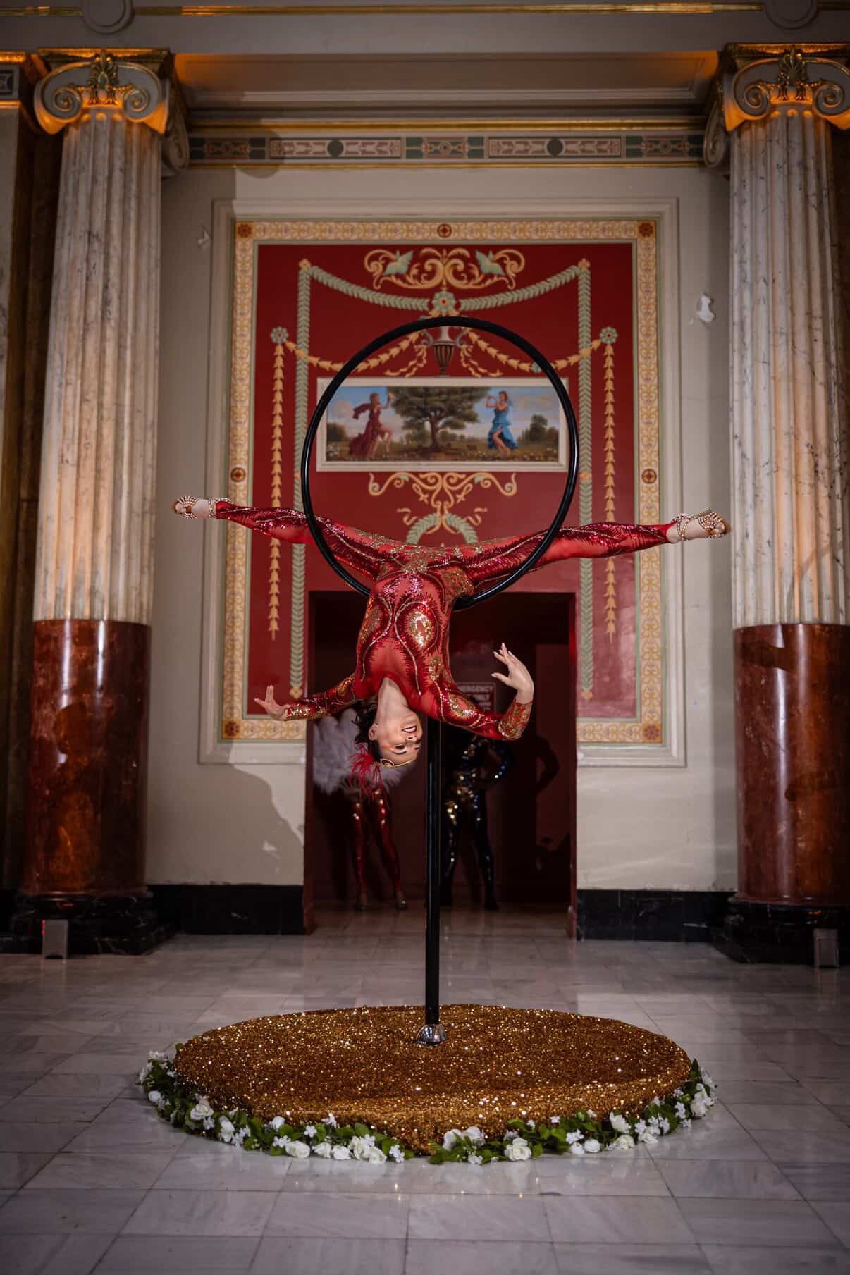 An acrobat in a custom-made black sequin bodysuit performs an upside-down split on a hoop in an ornate hall, wearing the Showgirl Costume, Sequin Bodysuit for Women or Men.