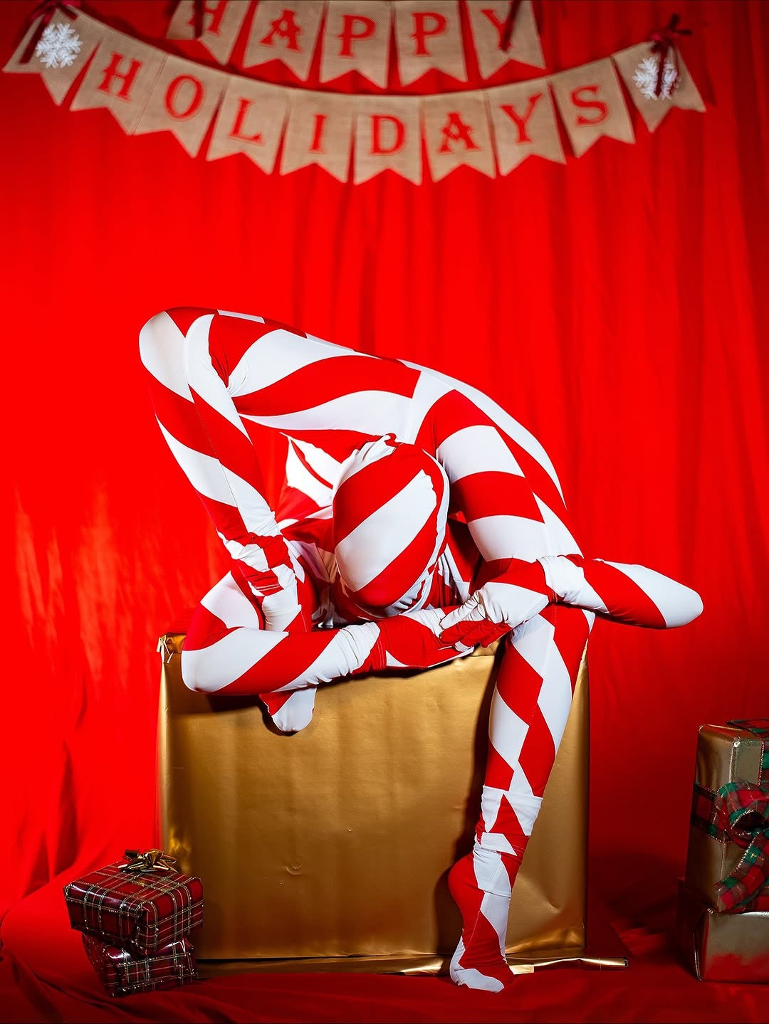 Wearing the Candy Cane Bodysuit turtleneck with full sleeves, someone poses on a gold gift box under a "Happy Holidays" banner.