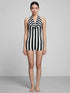 A woman stands barefoot against a gray background, wearing the Black and White Striped Leotard featuring a halter top with an open back.