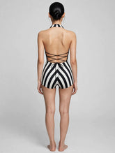 A woman stands barefoot with her back to the camera, wearing a black and white striped leotard featuring a halter top and open back.