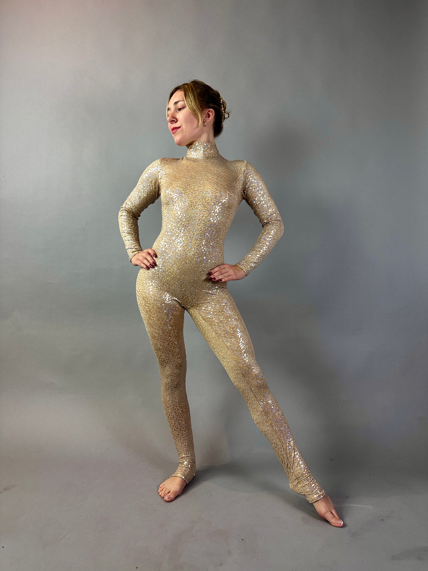 A woman poses confidently against a plain gray background in the Champagne Fairy Sequin Catsuit featuring an open back, turtleneck, and sleeves.