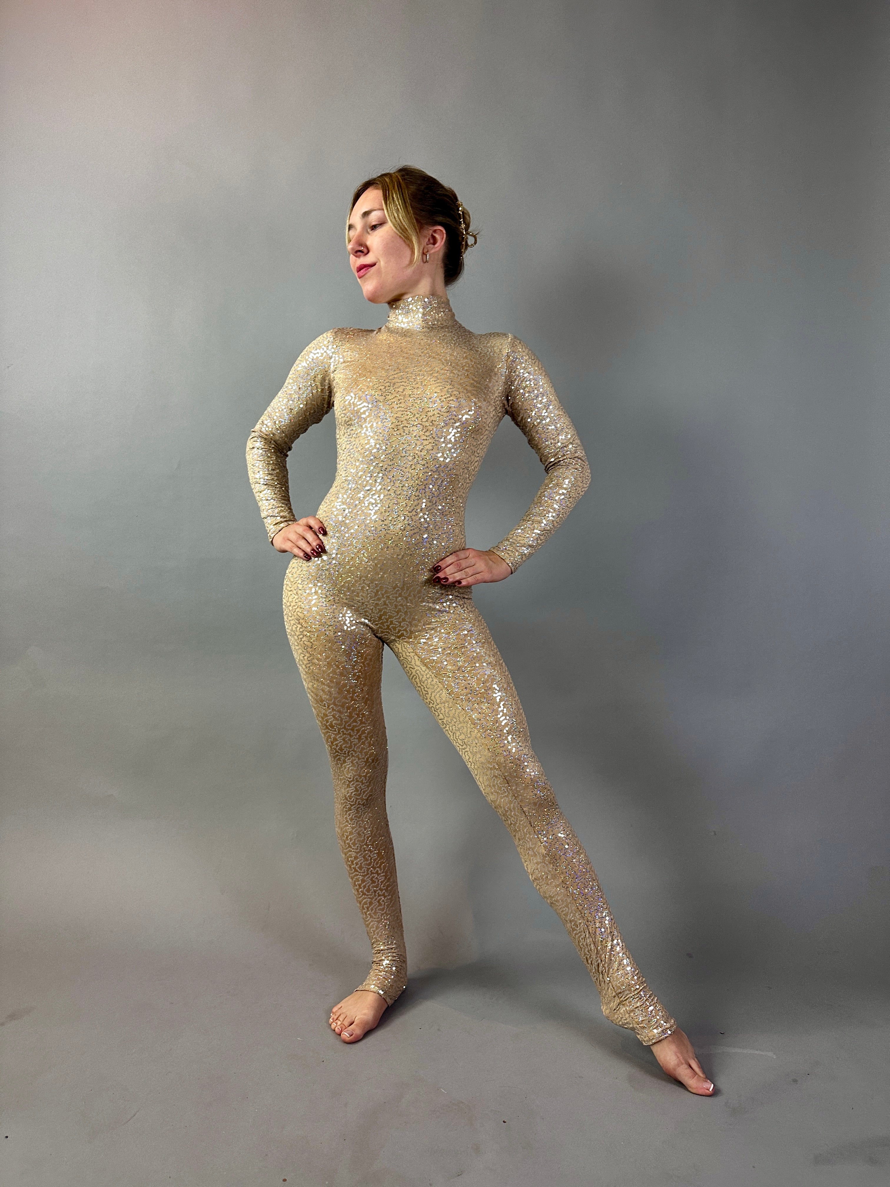 A woman poses confidently against a plain gray background in the Champagne Fairy Sequin Catsuit featuring an open back, turtleneck, and sleeves.
