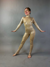 Barefoot, a person poses with arms outstretched in the Champagne Fairy Sequin Catsuit—its gold, sparkling fabric shimmering against a plain grey backdrop. The catsuit features an open back, turtleneck, and sleeves.