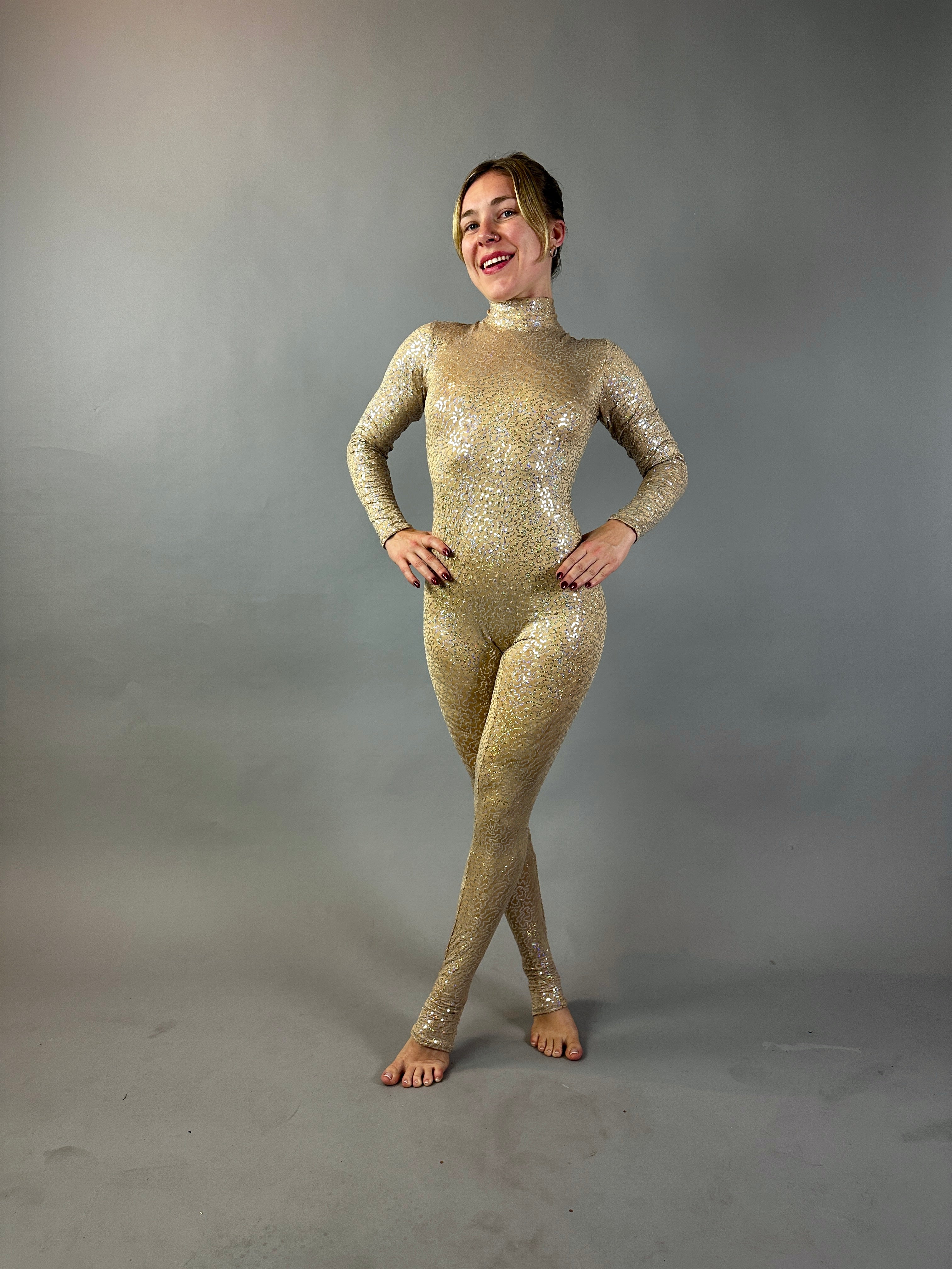 Model poses in the Champagne Fairy Sequin Catsuit featuring a turtleneck, sleeves, and open back. The sparkly gold iridescent sequins shine against a plain grey background as she smiles with hands on hips.