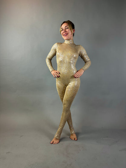 Model poses in the Champagne Fairy Sequin Catsuit featuring a turtleneck, sleeves, and open back. The sparkly gold iridescent sequins shine against a plain grey background as she smiles with hands on hips.