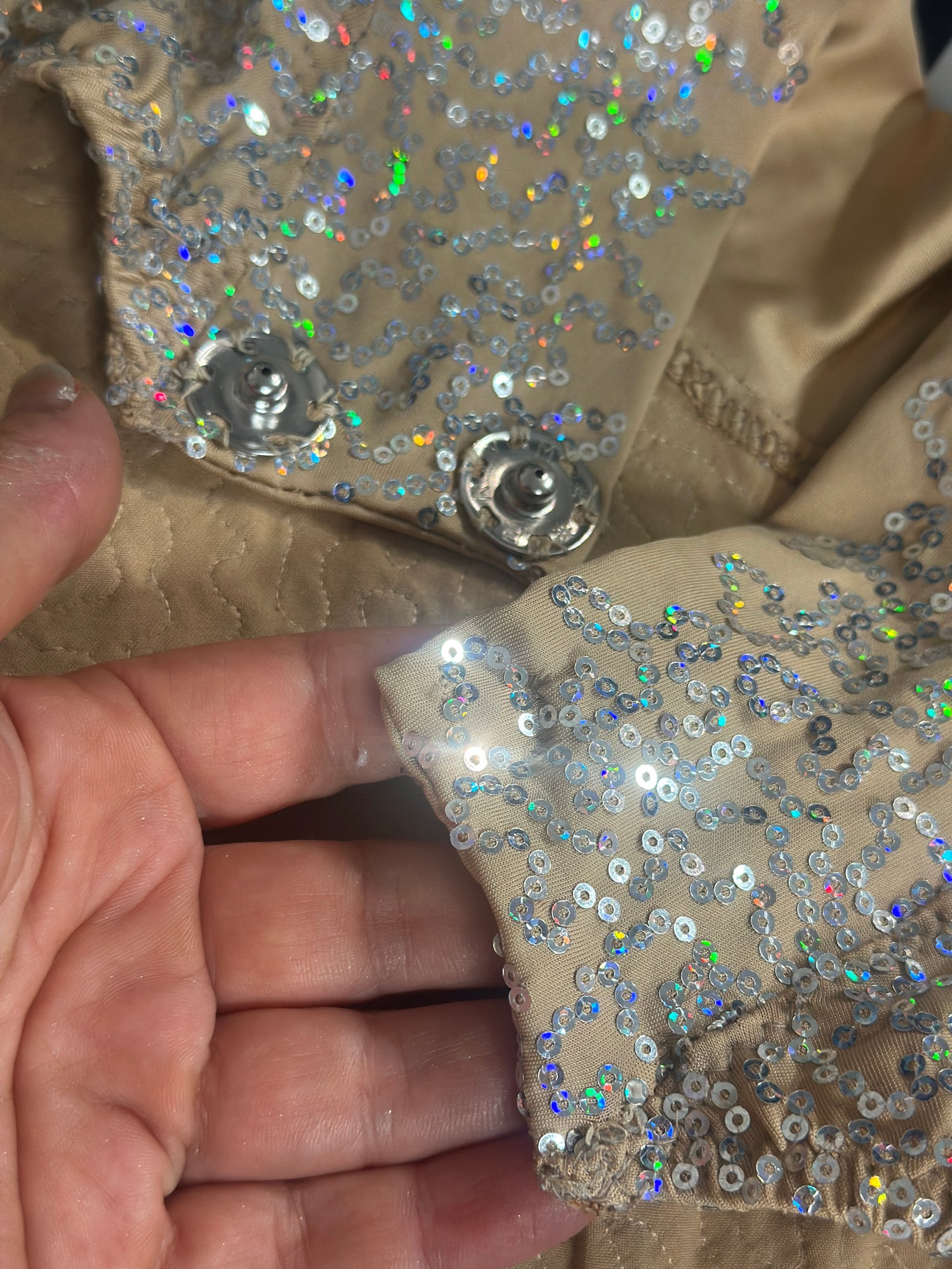 A hand holds the Champagne Fairy Sequin Catsuit’s beige fabric with shiny, iridescent sequins and a metal snap button—an open-back, turtleneck piece with sleeves, perfect as a dazzling performance costume.