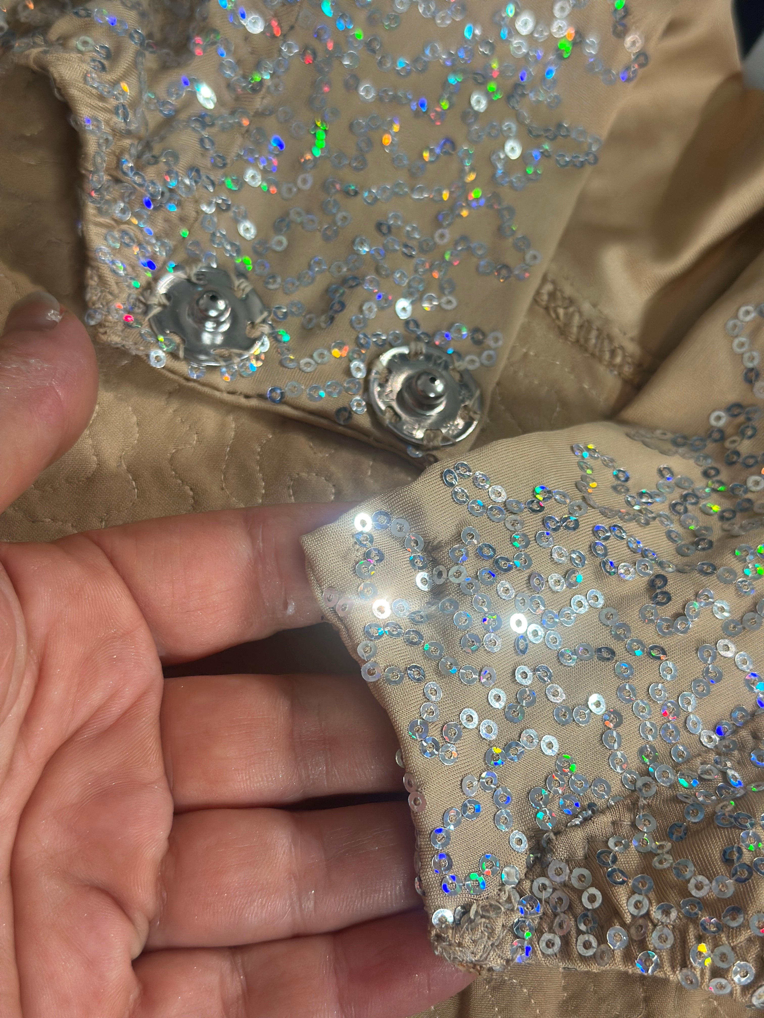 A hand holds the Champagne Fairy Sequin Catsuit’s beige fabric with shiny, iridescent sequins and a metal snap button—an open-back, turtleneck piece with sleeves, perfect as a dazzling performance costume.