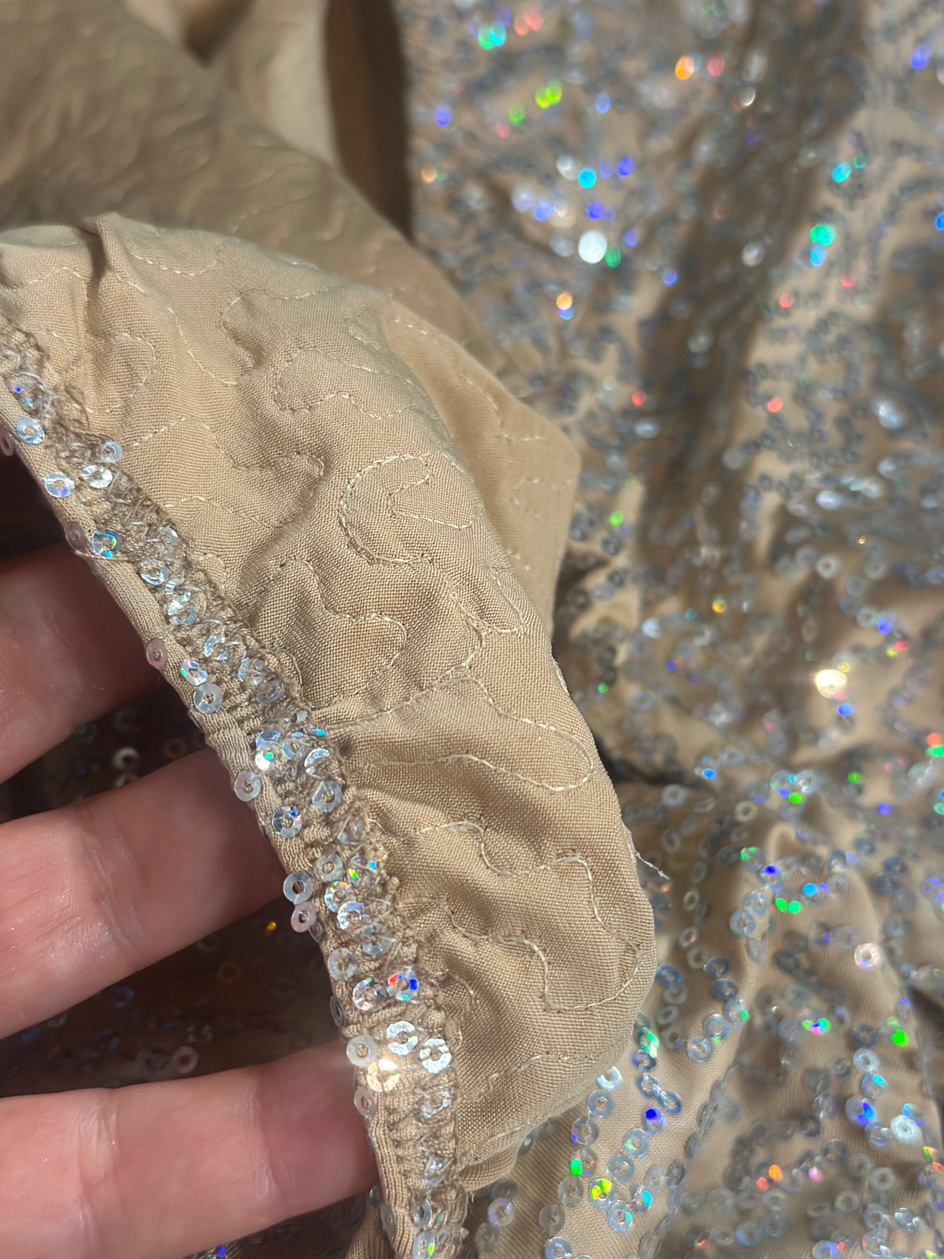 A hand displays beige fabric with silver sequins and holographic sparkles—ideal for creating the Champagne Fairy Sequin Catsuit, featuring an open back, turtleneck, and sleeves for a dazzling performance look.