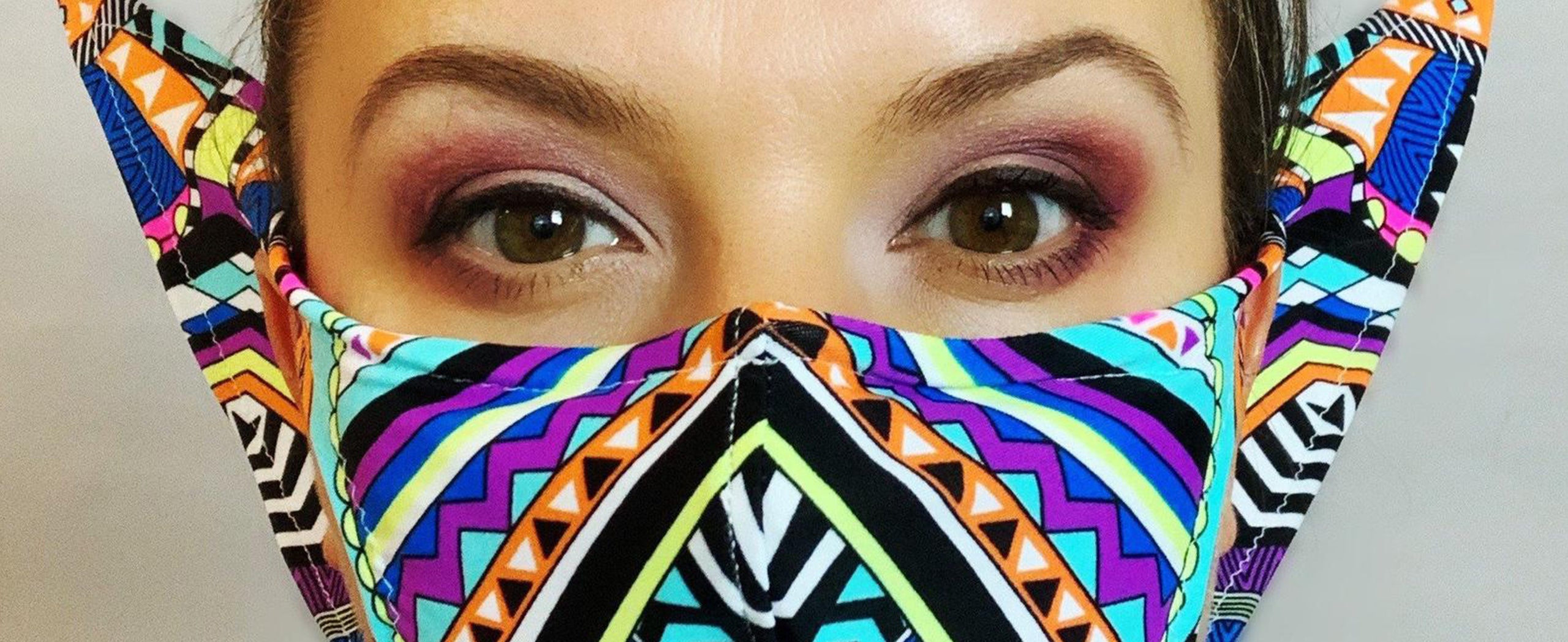 Person wearing a colorful, geometric-patterned face mask, revealing only their eyes, which are adorned with makeup.