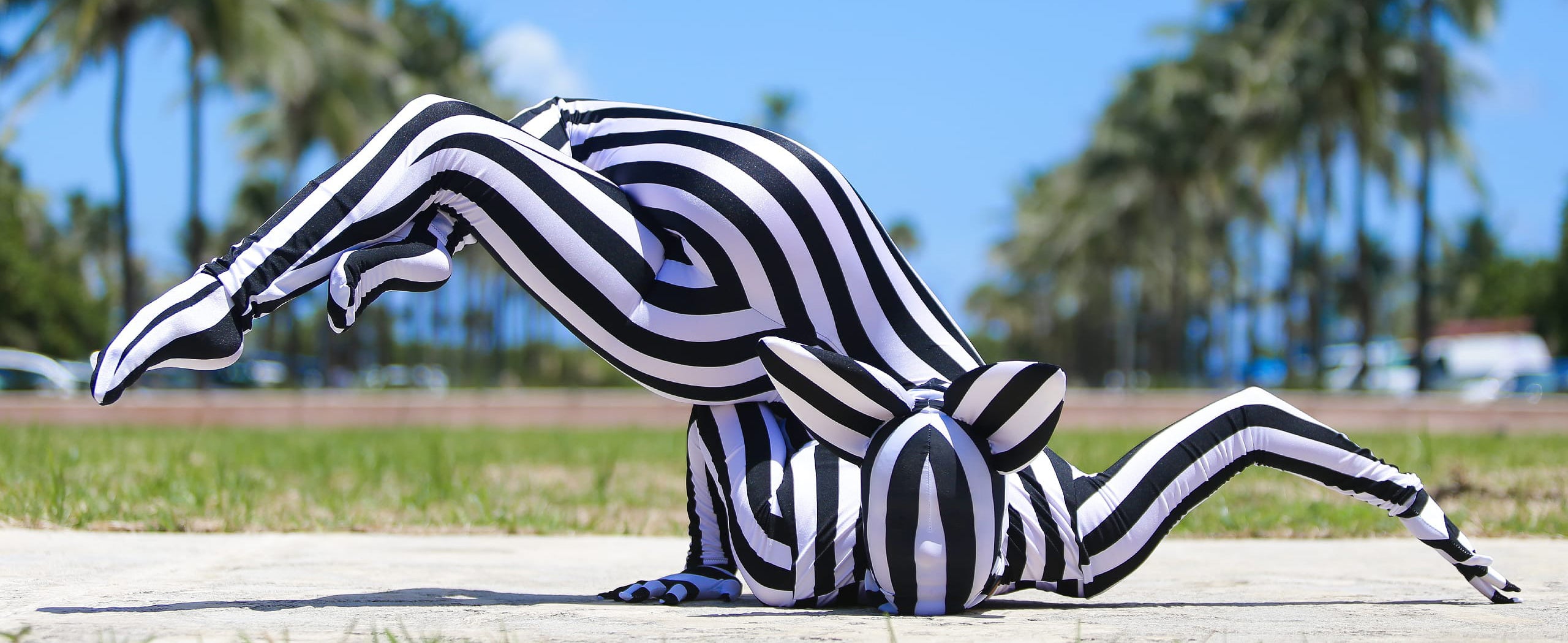 Person in a striped bodysuit performs a flexible pose on the ground with palm trees and a blue sky in the background.