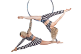 Two women wear asymmetrical harlequin catsuits—checkerboard spandex jumpsuits with one sleeve, one leg, and cutout design—as they perform an acrobatic pose on an aerial hoop against a white background.