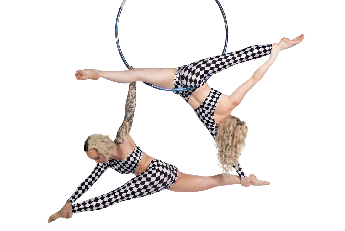 Two women wear asymmetrical harlequin catsuits—checkerboard spandex jumpsuits with one sleeve, one leg, and cutout design—as they perform an acrobatic pose on an aerial hoop against a white background.