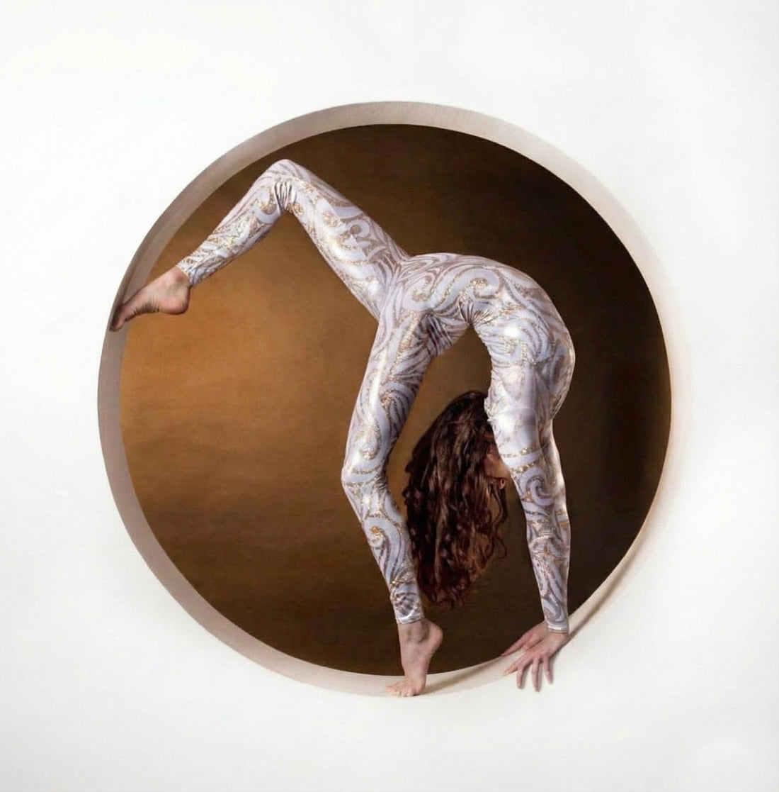 A woman wearing a White and Gold Catsuit Bodysuit with full sleeves and a round neck does a handstand inside a circular frame against a brown background.