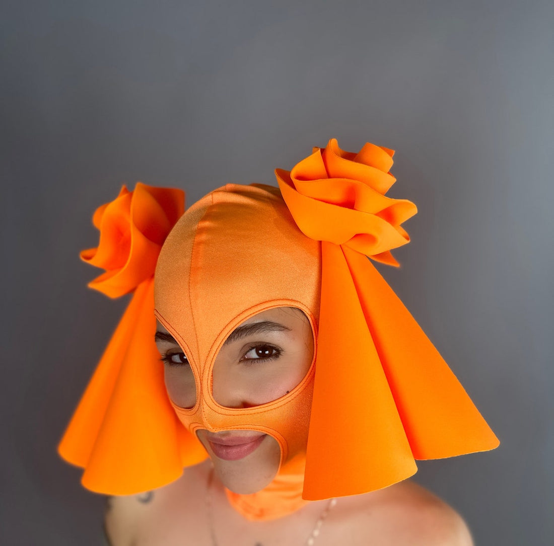 A person in an orange mask with floral embellishments and triangular side panels stands against a gray background, complemented by a glow-in-the-dark Circus Headpiece, perfect for fun costumes, exotic dance wear, clubbing, and festival or rave fashion.