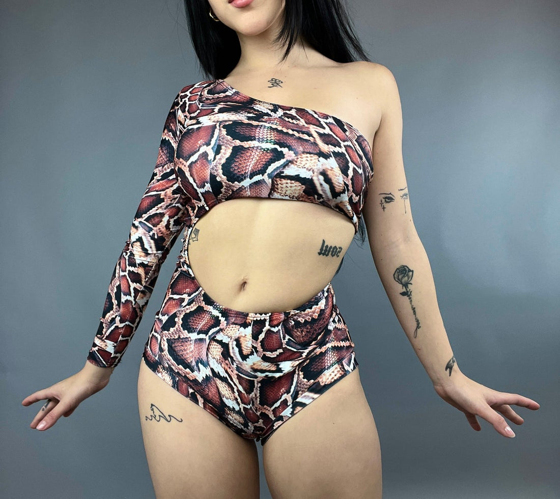 A person stands against a gray background wearing a one-sleeve snake print swimsuit that highlights their tattoos, reminiscent of the Leotard Costume for Gymnasts.