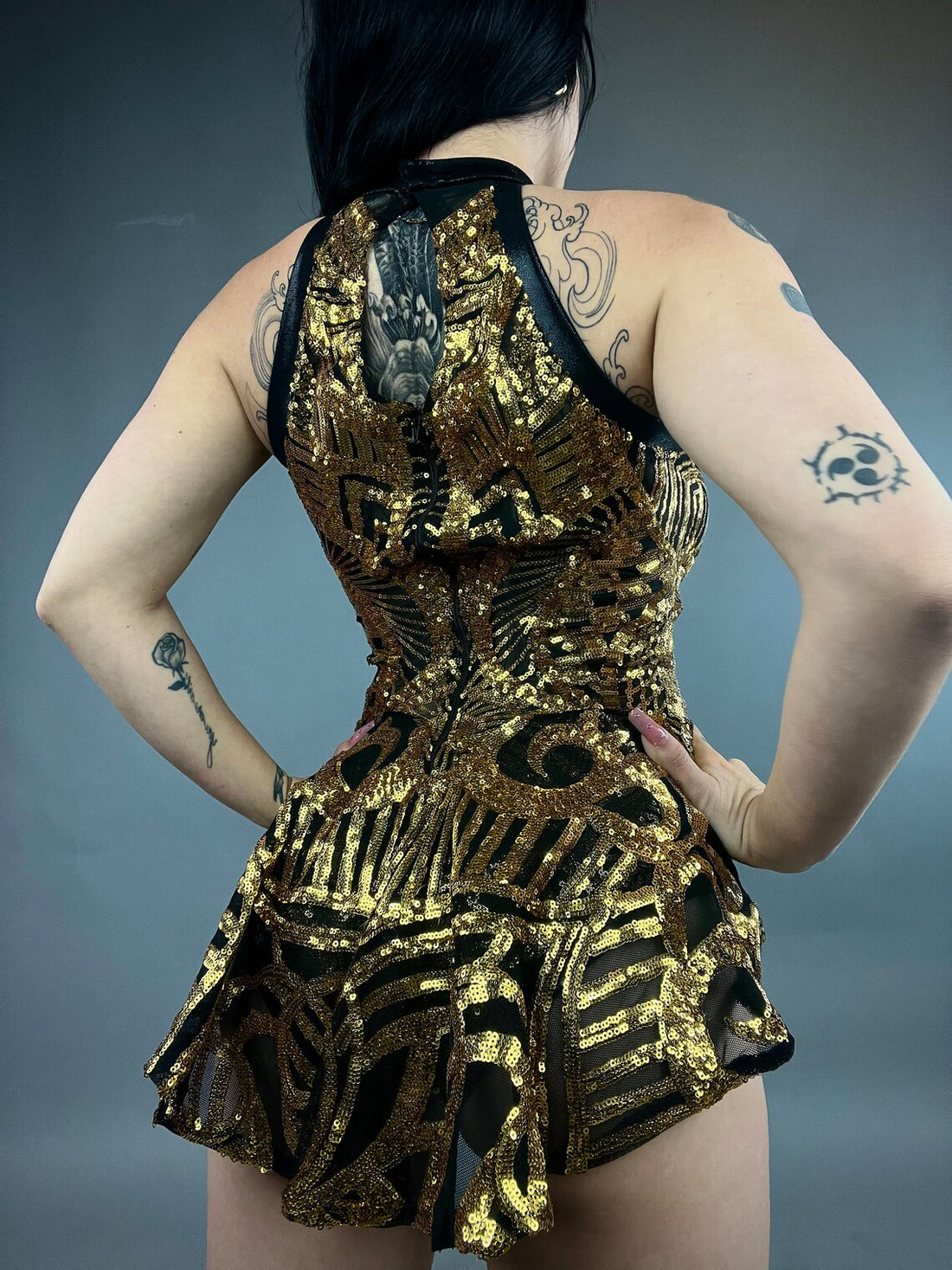 The person exudes confidence in the Show Girl Costume, a stunning black and gold sequin leotard with bold tattoos against a gray background—ideal for a showgirl spectacle or stage performance.