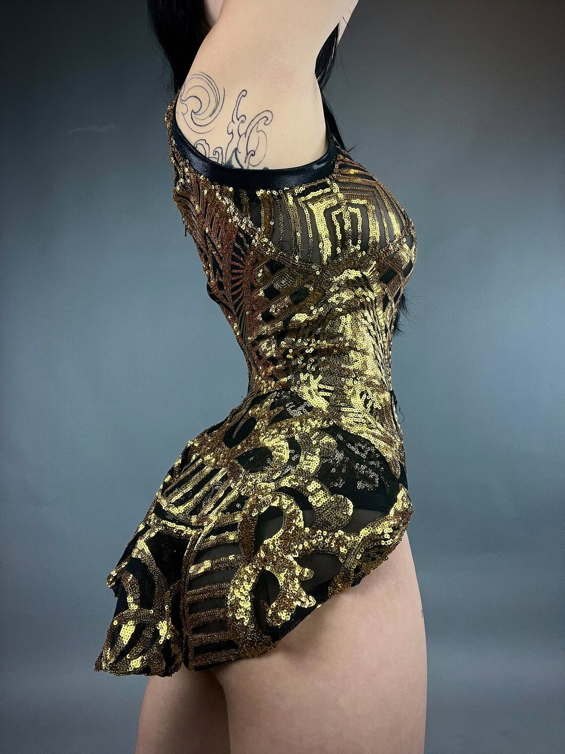 A tattooed woman poses against a plain backdrop wearing a Show Girl Costume, Black Gold Sequins Leotard, reminiscent of exotic dance wear, shimmering like sequin mesh under the spotlight.