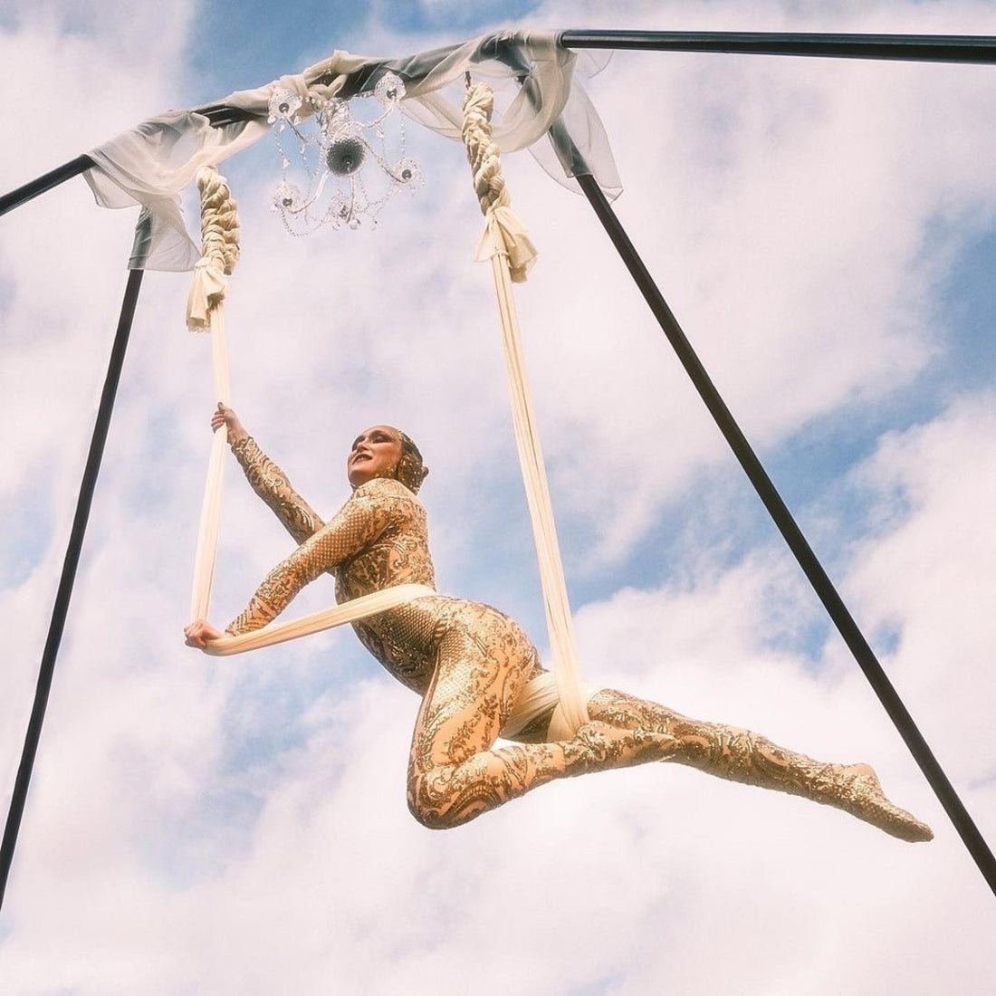 An aerial performer in a Gold Sequin Catsuit, part of the Gold Fairy Costume with full sleeves, gracefully swings from white silks against a cloudy sky.