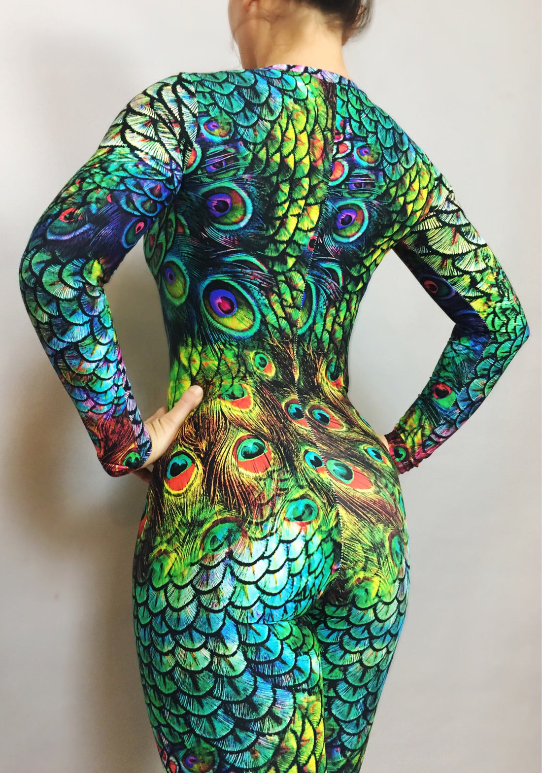 A person poses elegantly against a plain background in a full-sleeved, round-neck Peacock Print Spandex Catsuit. This custom-fit, limited edition piece beautifully captures the vibrant allure of a peacock’s plumage with exquisite detail.