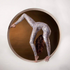 A contortionist in a white and gold catsuit bodysuit with full sleeves and round neck bends gracefully through a circular frame against a rich brown background.