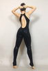 Barefoot and facing away, a person embodies futuristic fashion in the sleek Black Open Back Catsuit, featuring a halterneck and sleeveless design, with hands on their head.