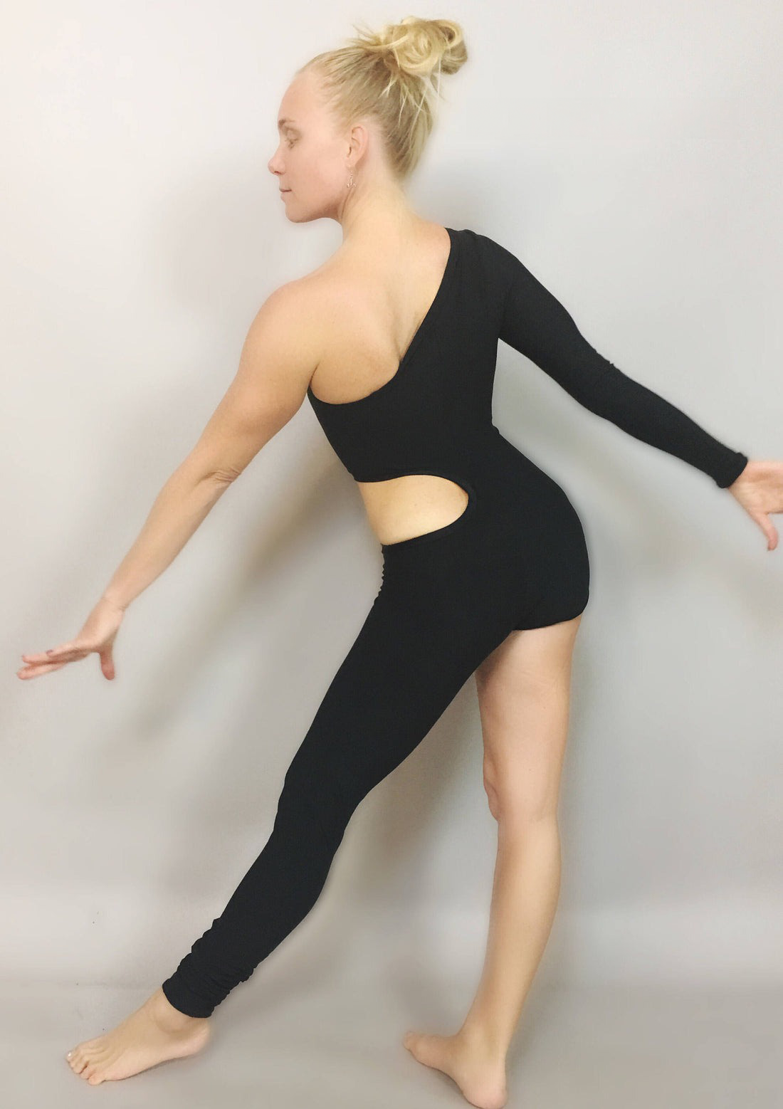 A person poses gracefully in a Unisex Isolated Black Catsuit with an asymmetric design and cut-outs, highlighting the elegance of performance wear against a plain backdrop.