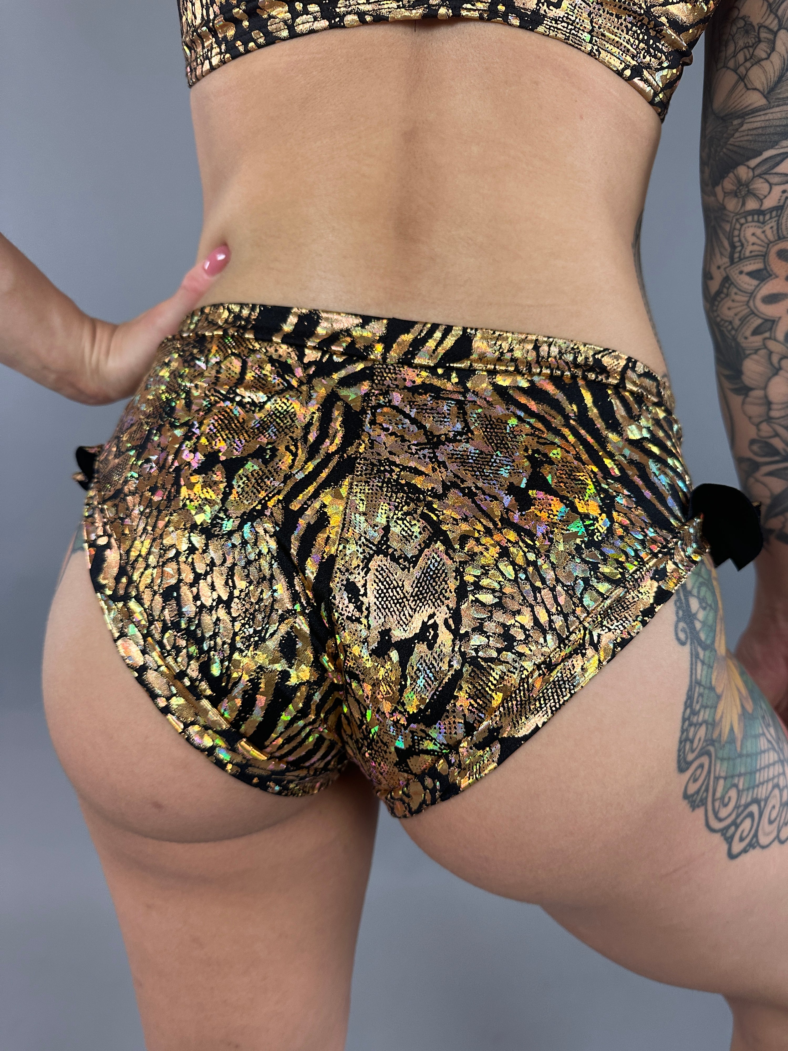 A person in Gold &amp; Black Snakeskin Crop Top &amp; Bottom Set, showcasing tiger and snake print shorts, stands against a gray background, highlighting their tattooed arm and leg.