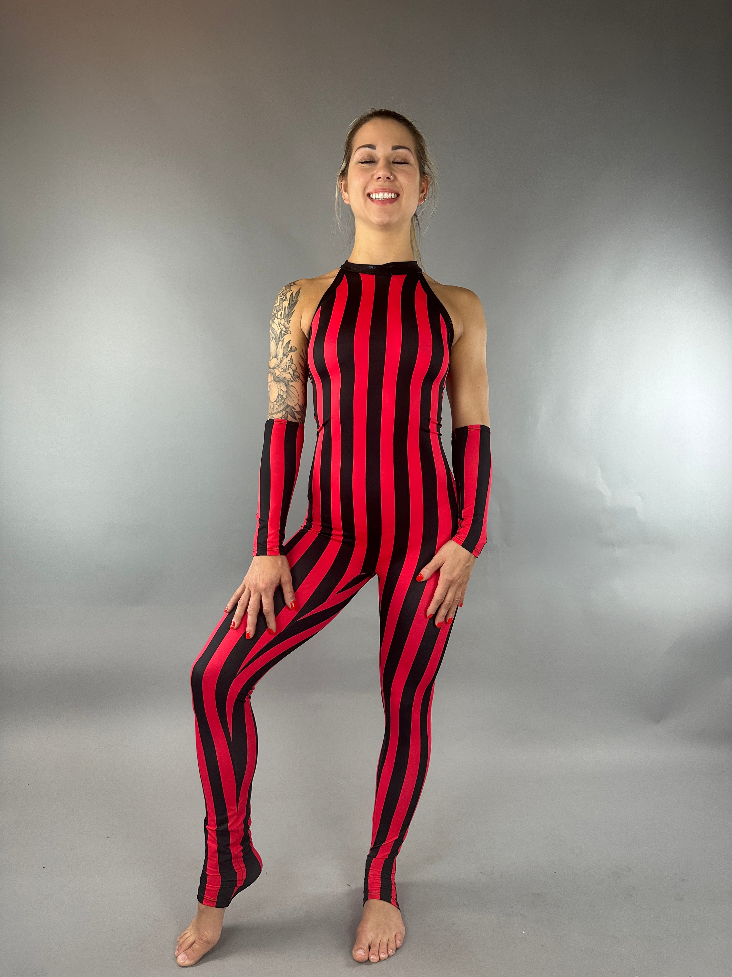A person in a circus costume, wearing the Red and Black Stripes Catsuit—a sleeveless halter neck jumpsuit with hand accessories—poses with a smile against a plain backdrop.