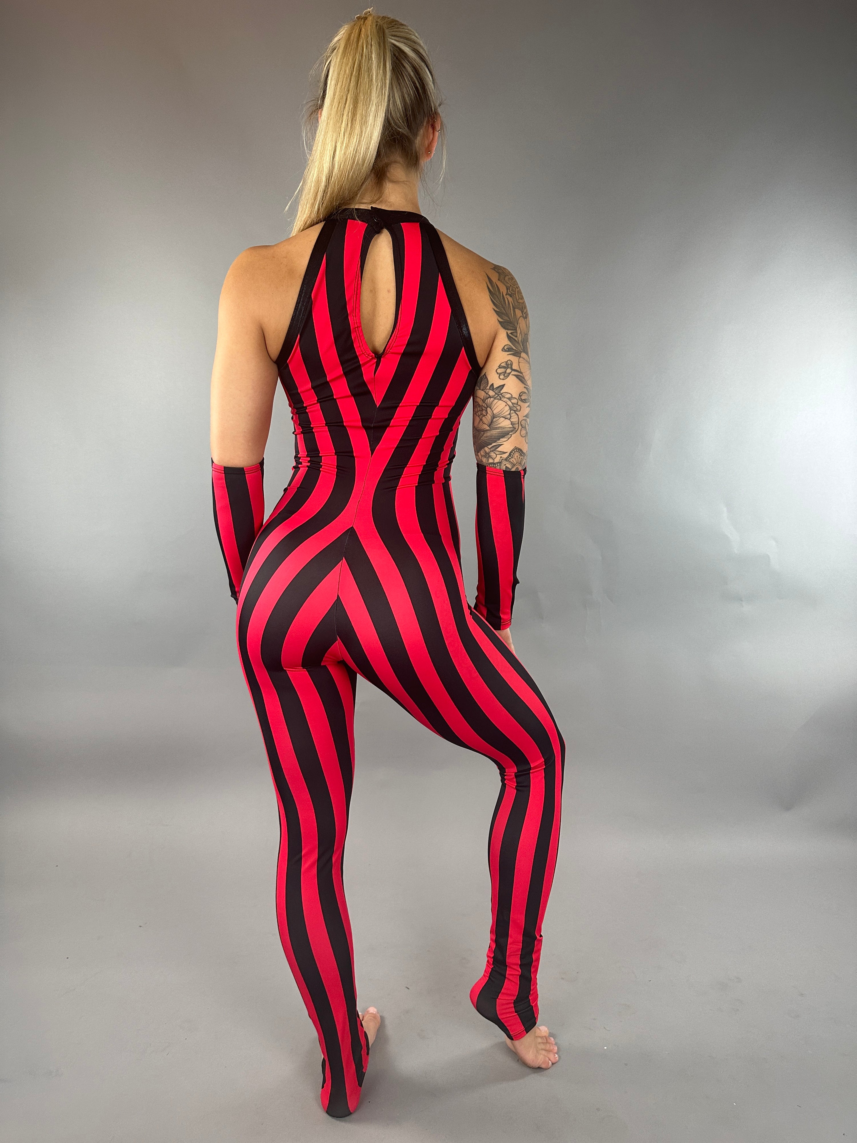 A person in a dazzling circus costume poses effortlessly against a gray background, wearing the Red and Black Stripes Catsuit, a sleeveless jumpsuit with a halter neck and hand accessories, evoking the grace of aerialists and acrobats.