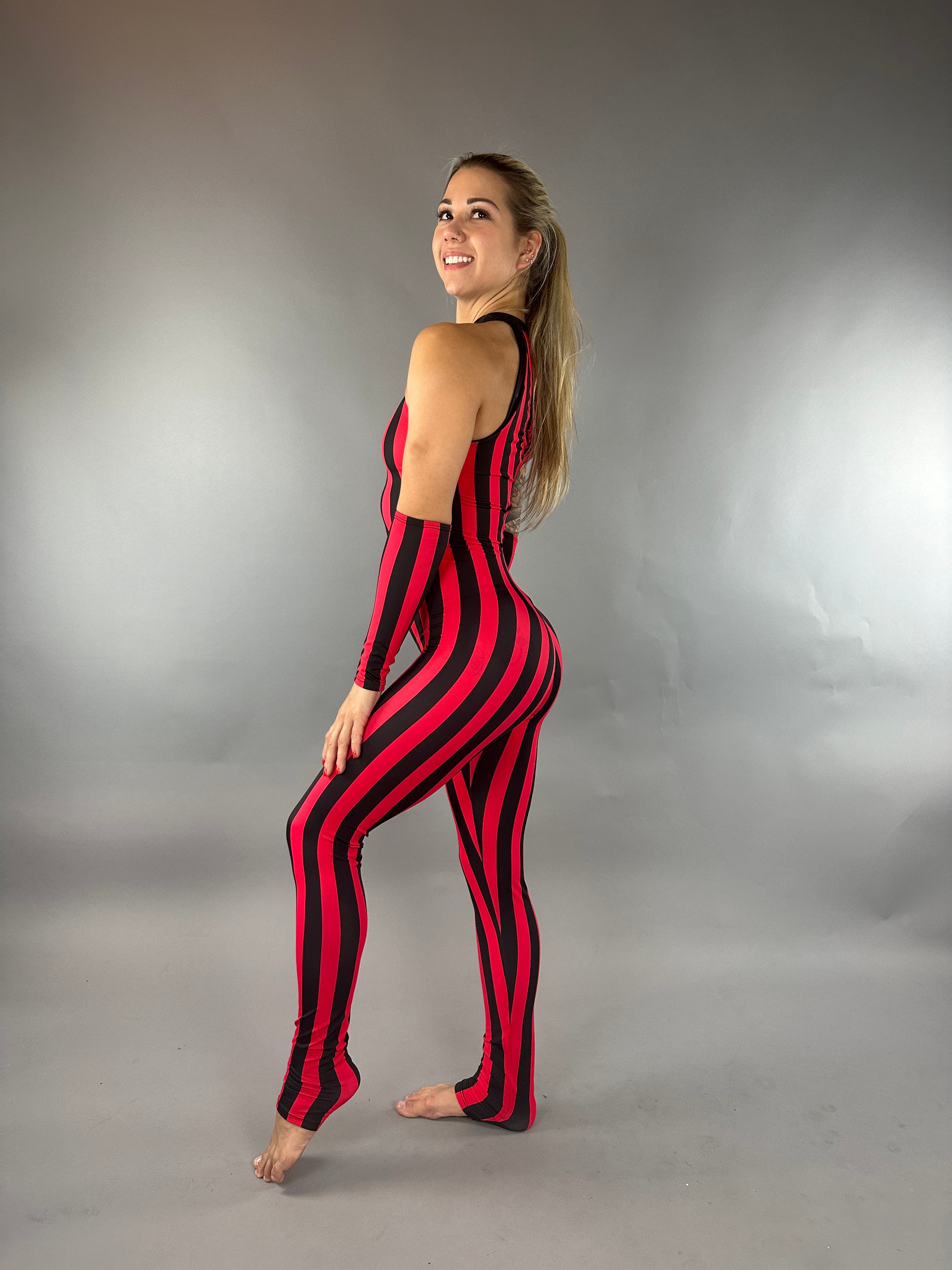 A woman in a Red and Black Stripes Catsuit, a sleeveless jumpsuit reminiscent of a circus costume, is smiling and posing gracefully against a plain gray background.