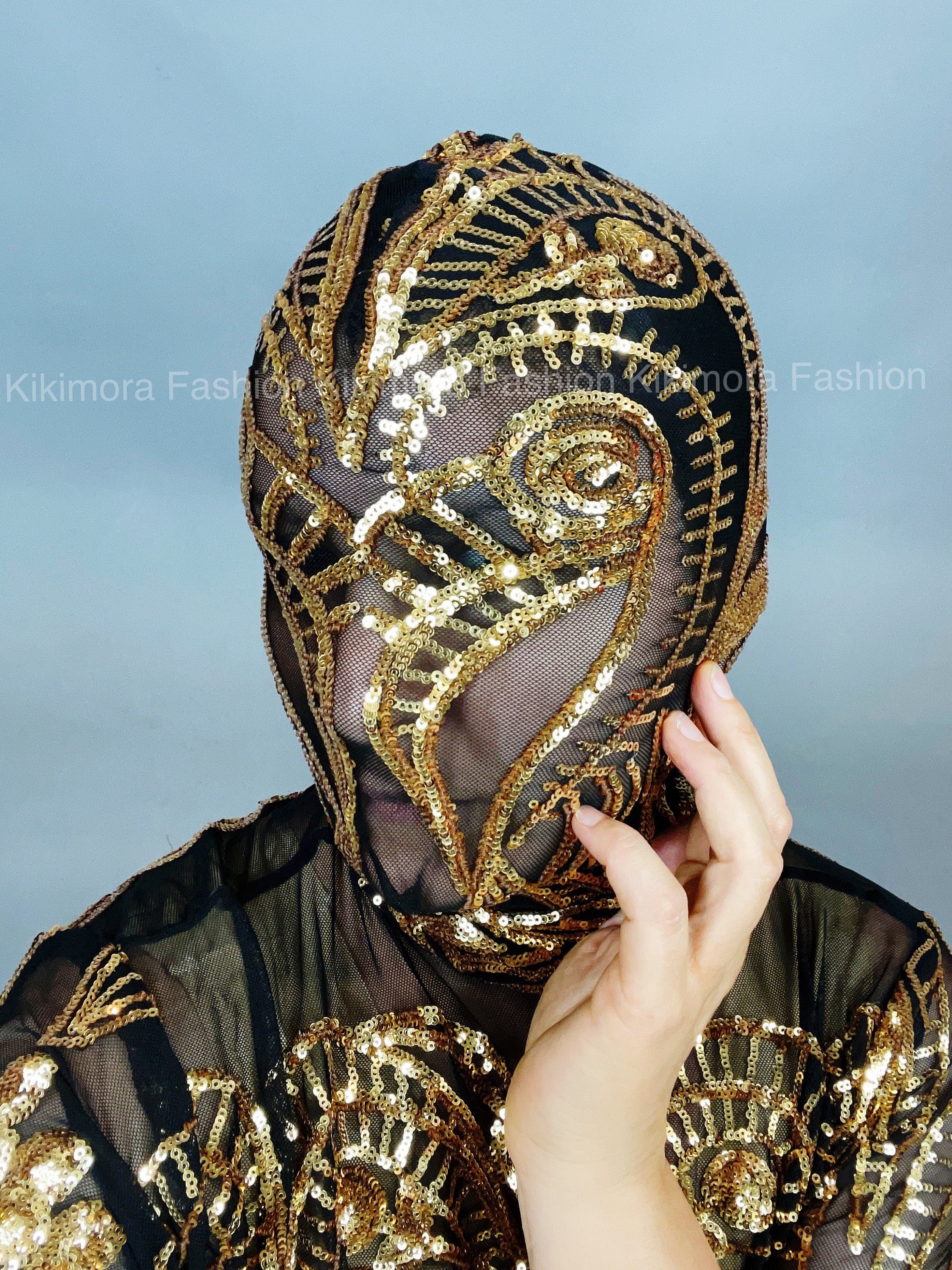 A person in a Gold Sequin on Black Sheer Dress with thigh-high slits and a turtleneck is touching their face, wearing an ornate sequined mask.