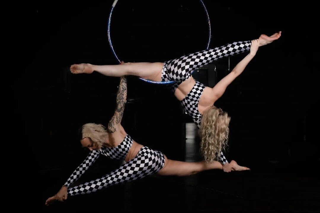 Two acrobats perform a balancing act in Harlequin Costume Spandex Jumpsuits, one supporting the other against a dark background.