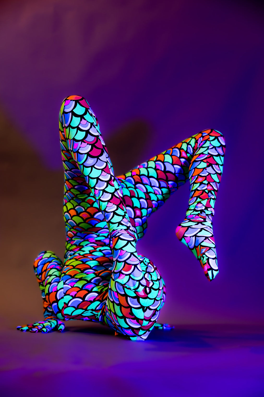 Against a purple backdrop, someone in a vibrant scale-patterned bodysuit poses flexibly, capturing the essence of the Neon Mermaid Alien Costume, Full Body Catsuit with Headpiece.