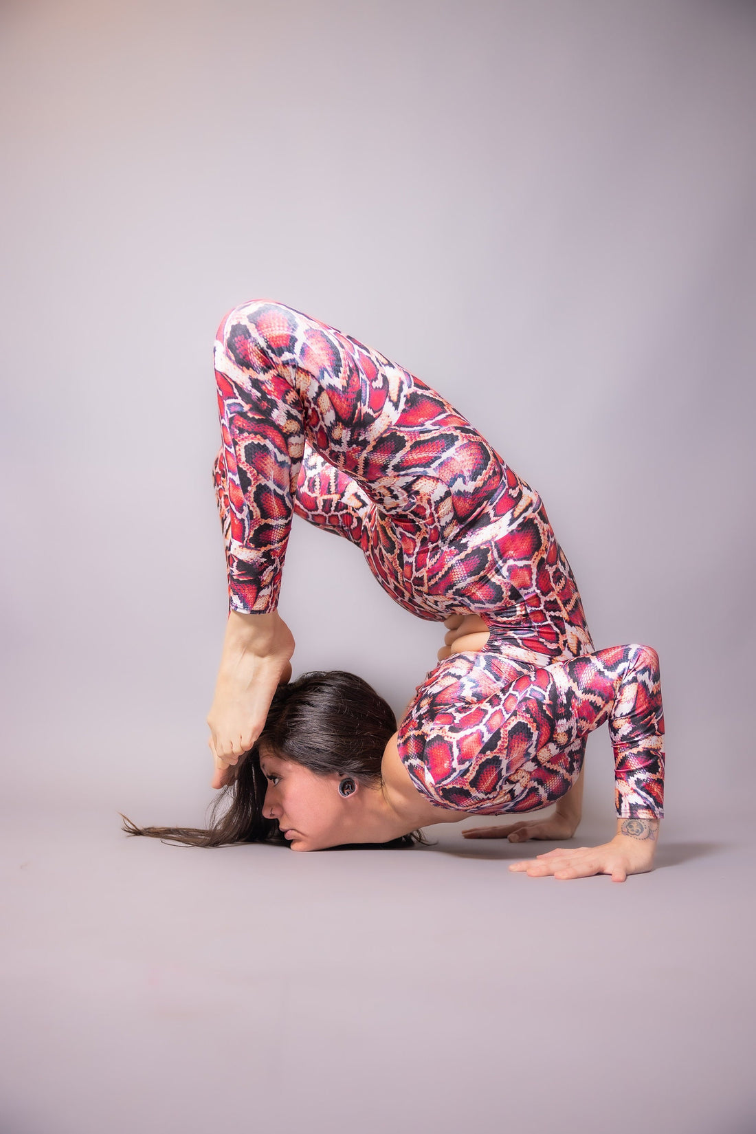 Someone wearing the Red Snake-Print Isolated Catsuit, featuring a waist cutout, effortlessly performs an advanced yoga pose by bending backward with feet over their head, showcasing the flexibility and vibrancy of this stunning Lycra jumpsuit.